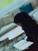 Young traditional muslim woman reading Quran on the sofa before iftar dinner during a ramadan feast at home photo