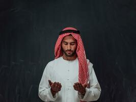 Arabian man in traditional clothes making traditional prayer to God, keeps hands in praying gesture in front of black chalkboard representing modern islam fashion and ramadan kareem concept photo