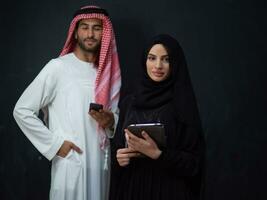 Young muslim business couple arabian man with woman in fashionable hijab dress using mobile phone and tablet computer in front of black chalkboard representing modern islam fashion technology photo