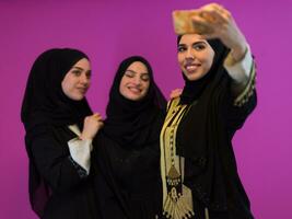 Group of beautiful muslim women two of them in fashionable dress with hijab using mobile phone while taking selfie picture isolated on pink background representing modern islam fashion technology photo