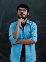 un joven indio hombre en un azul camisa y lentes poses pensativamente en frente de colegio tablero foto