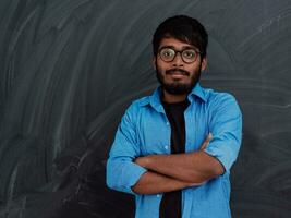 A young Indian student in a blue shirt with glasses posing with his arms crossed in front of the school blackboard photo