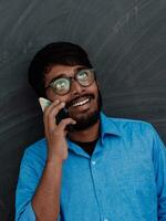 Indian smiling young student in blue shirt and glasses using smartphone and posing on school blackboard background photo