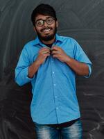 Indian young student wearing glasses puts on a blue shirt while standing in front of the school blackboard photo