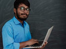 Indian smiling young student in blue shirt and glasses using laptop and posing on school blackboard background photo