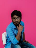 A young Indian student in a blue shirt with glasses sitting in front of a pink background photo