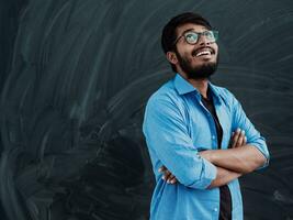 A young Indian student in a blue shirt with glasses posing with his arms crossed in front of the school blackboard photo