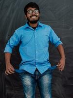 Photo of an Indian man wearing a blue shirt and glasses while symbolically showing his empty pockets on the background of a school blackboard.