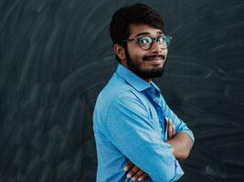 A young Indian student in a blue shirt with glasses posing with his arms crossed in front of the school blackboard photo