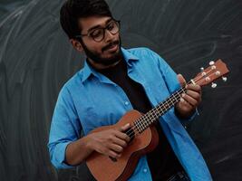 Indian young man in a blue shirt and glasses playing the guitar in front of the school blackboard photo