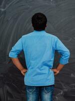 A young Indian student in a blue shirt with his back to the camera is standing in front of the school blackboard photo