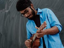 Indian young man in a blue shirt and glasses playing the guitar in front of the school blackboard photo
