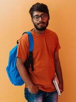 indio estudiante con azul mochila, lentes y cuaderno posando en naranja antecedentes. el concepto de educación y enseñanza. hora a Vamos espalda a colegio foto