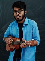 Indian young man in a blue shirt and glasses playing the guitar in front of the school blackboard photo