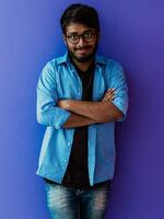 A young Indian student in a blue shirt with glasses posing with his arms crossed posing on purple photo