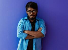 un joven indio estudiante en un azul camisa con lentes posando con su brazos cruzado posando en púrpura foto