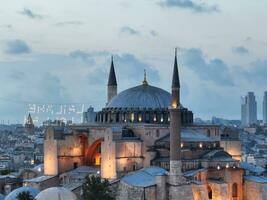 Establishing orbiting aerial drone shot of a Hagia Sophia Holy Grand Mosque with Bosphorus bridge and city skyline with a flag on the background in Fatih, Istanbul, Turkey at sunset. photo