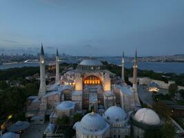 Establishing orbiting aerial drone shot of a Hagia Sophia Holy Grand Mosque with Bosphorus bridge and city skyline with a flag on the background in Fatih, Istanbul, Turkey at sunset. photo