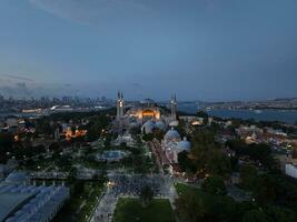 Establishing orbiting aerial drone shot of a Hagia Sophia Holy Grand Mosque with Bosphorus bridge and city skyline with a flag on the background in Fatih, Istanbul, Turkey at sunset. photo