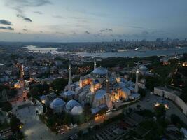 Establishing orbiting aerial drone shot of a Hagia Sophia Holy Grand Mosque with Bosphorus bridge and city skyline with a flag on the background in Fatih, Istanbul, Turkey at sunset. photo