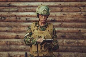 Woman soldier using tablet computer against old wooden wall in camp photo