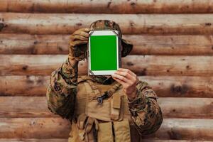 soldado utilizando tableta computadora en contra antiguo de madera pared en militar acampar a permanecer en contacto con amigos y familia foto