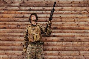 A soldier in uniform with a rifle in his hand is standing in front of a wooden wall. A soldier guards the forest base from the enemy photo
