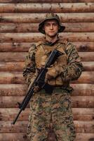 un soldado en uniforme con un rifle en su mano es en pie en frente de un de madera pared. un soldado guardias el bosque base desde el enemigo foto