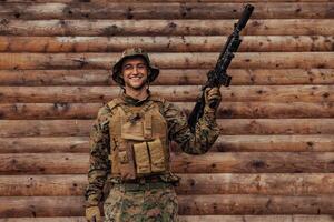 A soldier in uniform with a rifle in his hand is standing in front of a wooden wall. A soldier guards the forest base from the enemy photo