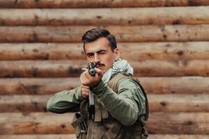 A soldier in uniform with a rifle in his hand is standing in front of a wooden wall. A soldier guards the forest base from the enemy photo