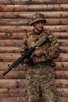 A soldier in uniform with a rifle in his hand is standing in front of a wooden wall. A soldier guards the forest base from the enemy photo