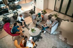 Top view photo of Multiethnic startup business team having brainstorming in relaxation area of modern office interior working on laptop and tablet computer