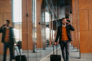 ir a la terminal del aeropuerto. viajero de negocios confiado caminando por las calles de la ciudad y tirando de su maleta bebiendo café y hablando en el teléfono inteligente foto