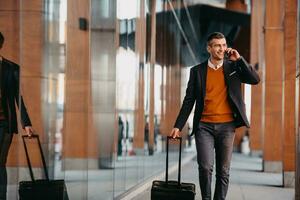 ir a la terminal del aeropuerto. viajero de negocios confiado caminando por las calles de la ciudad y tirando de su maleta bebiendo café y hablando en el teléfono inteligente foto