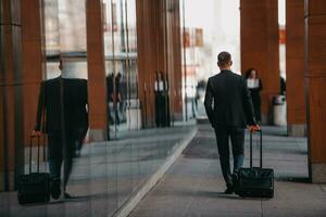 Going to airport terminal. Confident businessman traveler walking on city streets and pulling his suitcase drinking coffee and speaking on smartphone photo