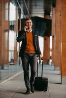 Going to airport terminal. Confident businessman traveler walking on city streets and pulling his suitcase drinking coffee and speaking on smartphone photo