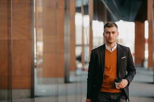 Going to airport terminal. Confident businessman traveler walking on city streets and pulling his suitcase drinking coffee and speaking on smartphone photo