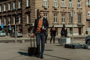 Going to airport terminal. Confident businessman traveler walking on city streets and pulling his suitcase drinking coffee and speaking on smartphone photo