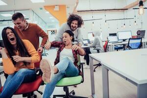 Team building and office fun. Young cheerful businesspeople in smart casual wear having fun while racing on office chairs and smiling. photo