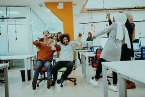 Team building and office fun. Young cheerful businesspeople in smart casual wear having fun while racing on office chairs and smiling. photo