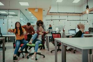 Team building and office fun. Young cheerful businesspeople in smart casual wear having fun while racing on office chairs and smiling. photo