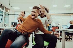 Team building and office fun. Young cheerful businesspeople in smart casual wear having fun while racing on office chairs and smiling. photo