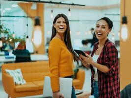 Portrait of two businesswomen talking to each other while standing in a modern business office with their colleagues using laptop and tablet.. Marketing concept. Multi-ethnic society. photo