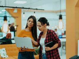 Portrait of two businesswomen talking to each other while standing in a modern business office with their colleagues using laptop and tablet.. Marketing concept. Multi-ethnic society. photo