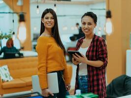 retrato de dos mujer de negocios hablando a cada otro mientras en pie en un moderno negocio oficina con su colegas utilizando ordenador portátil y tableta.. márketing concepto. multiétnico sociedad. foto