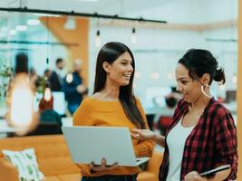 Portrait of two businesswomen talking to each other while standing in a modern business office with their colleagues using laptop and tablet.. Marketing concept. Multi-ethnic society. photo