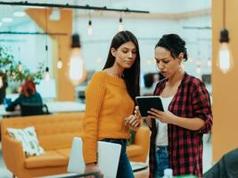 Portrait of two businesswomen talking to each other while standing in a modern business office with their colleagues using laptop and tablet.. Marketing concept. Multi-ethnic society. photo