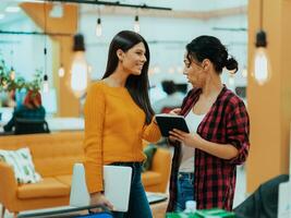 Portrait of two businesswomen talking to each other while standing in a modern business office with their colleagues using laptop and tablet.. Marketing concept. Multi-ethnic society. photo