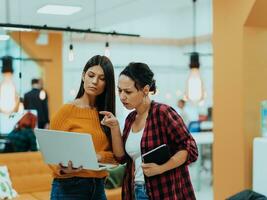 Portrait of two businesswomen talking to each other while standing in a modern business office with their colleagues using laptop and tablet.. Marketing concept. Multi-ethnic society. photo