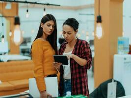Portrait of two businesswomen talking to each other while standing in a modern business office with their colleagues using laptop and tablet.. Marketing concept. Multi-ethnic society. photo